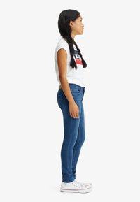 Jeans ajustados de denim azul con una textura suave, combinados con una camiseta de manga corta blanca con texto gráfico en rojo y negro, junto con zapatillas blancas.