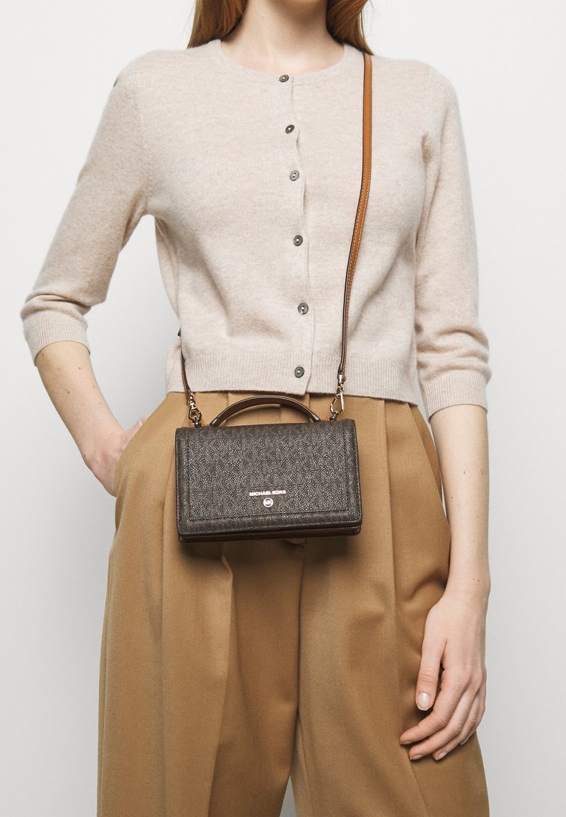 Ruskea kuvioitu käsilaukku, jossa on yläkahva ja irrotettava olkahihna, varustettuna logolla. Päällä beigepaitainen takki ja vaaleanruskeat housut.