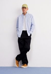 Une chemise boutonnée bleu clair et blanche à rayures, portée par-dessus un t-shirt blanc, associée à un pantalon noir et des baskets marron. Une casquette vert clair complète la tenue.