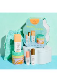 Daytime Exhibit skincare set with serum, eye cream, and anti-pollution drops, colorful packaging, and green patterned drawstring bag on blue surface.