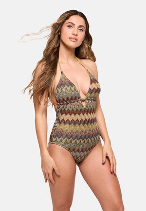 Mujer con cabello largo y ondulado, vestida con un bañador de una pieza con estampado y cuello halter, posando sobre un fondo liso y claro.