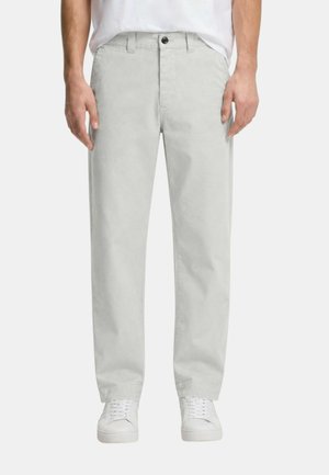 Mann trägt hellgraue Hose mit geradem Bein, Knopf und Reißverschluss, kombiniert mit weißen Sneakers und einem teilweise sichtbaren weißen Hemd.