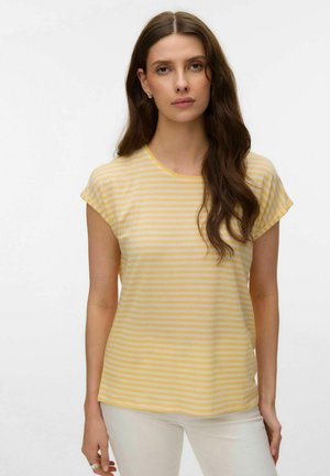 Woman with long brown hair wearing yellow and white striped short-sleeve shirt and light-colored pants, standing against a plain background.