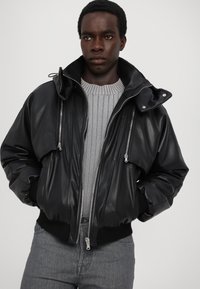 Veste bomber en simili cuir noir avec un col haut, une double fermeture éclair, des poignets côtelés et deux poches avant. Portée sur un pull gris texturé.