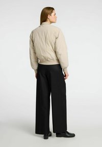 Una persona si trova con la schiena rivolta verso l'osservatore, indossando un giubbotto imbottito beige e pantaloni larghi neri, su uno sfondo chiaro.