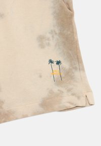 T-shirt en coton beige avec un motif tie-dye. Présente une broderie de palmier vert et le texte "PALM SWINGS" en jaune. Comprend des fentes sur les côtés.