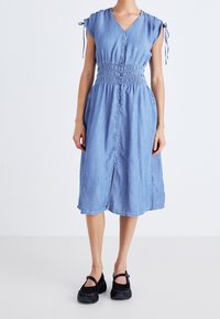 Nainen, jolla on päällään polvipituinen vaaleansininen denimitunika, jonka etuosassa on napit, rypytetty vyötärö, sidottavat hihansuut ja mustat Mary Jane -kengät.