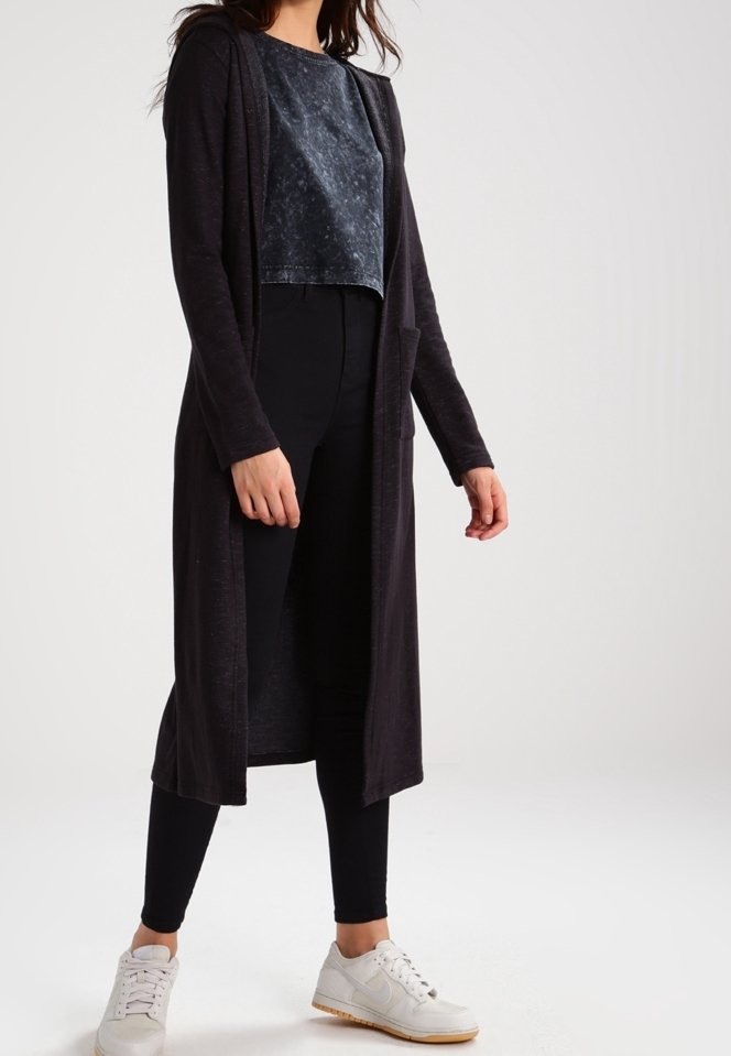 Femme portant un long cardigan noir, un top court noir délavé, un pantalon noir ajusté et des baskets blanches, debout sur un fond uni.