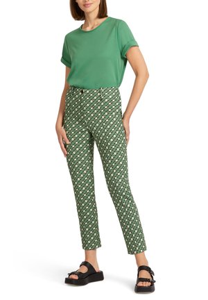 Femme debout, une main dans la poche d'un pantalon vert à motif, portant un haut vert uni à manches courtes et des sandales à plateforme noires sur fond blanc.