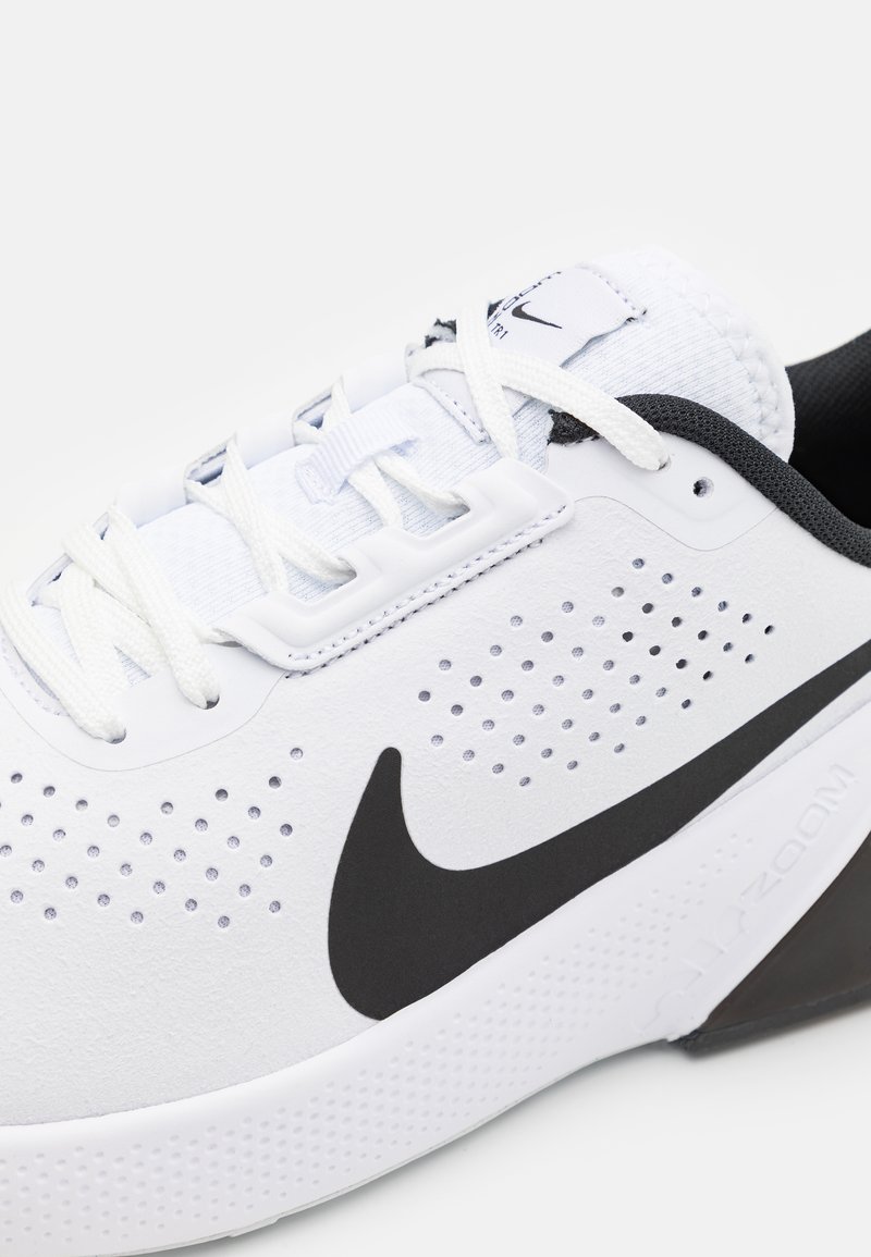 Zapatilla deportiva blanca con un logo negro, diseño perforado para ventilación, suela texturizada y cordones planos blancos.