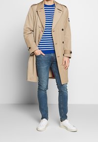 Béžový trenčkot přes modro-bíle pruhovaný svetr, doplněný modrými džínami a bílými teniskami. Kabát má dvouřadé zapínání.