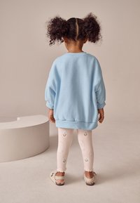Hellblaues oversized Sweatshirt, weiße Leggings mit kleinen Blumenmustern und flauschige Sandalen. Der Hintergrund ist in einem sanften neutralen Farbton gehalten.