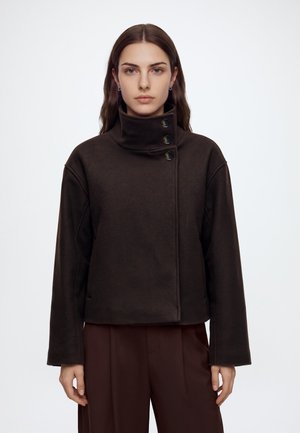 Femme aux longs cheveux bruns portant une veste marron foncé à col montant boutonnée et un pantalon marron assorti, faisant face à l'avant avec une expression neutre.