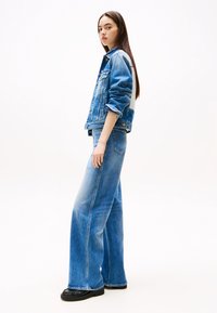 Heledad denim jakk ja laiad sinised teksad, mis on kergelt kulunud ning kontrastsete õmblustega. Mustad jalanõud täiendavad ansamblit.