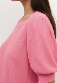 Roze gebreide top met korte pofmouwen, met een geribbelde textuur en een ronde halslijn. De stof lijkt zacht en lichtgewicht.