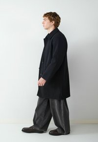 Schwarzer Mantel mit strukturiertem Design, ausgestattet mit einem Kragen und Knöpfen, kombiniert mit weiten grauen Jeans und dunklen Schuhen. Seitenansicht.