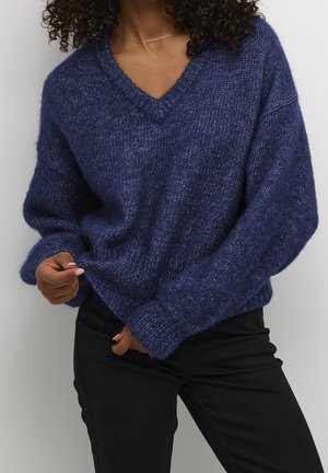 Kvinde iført en løs, tekstureret mørkeblå V-hals sweater og sorte bukser, justerer sweateren ved taljen med den ene hånd.