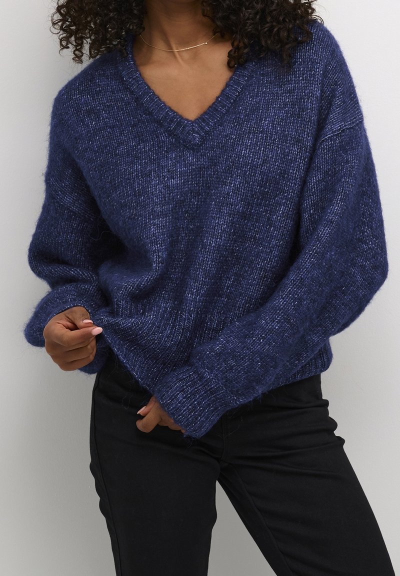 Femme portant un pull ample texturé bleu marine à col en V et un pantalon noir, ajustant le pull à la taille avec une main.