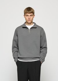 Jonge man met kort lichtbruin haar draagt een grijze sweatshirt met kwartrits, een wit shirt eronder en zwarte broek, staand met zijn handen in zijn zakken.