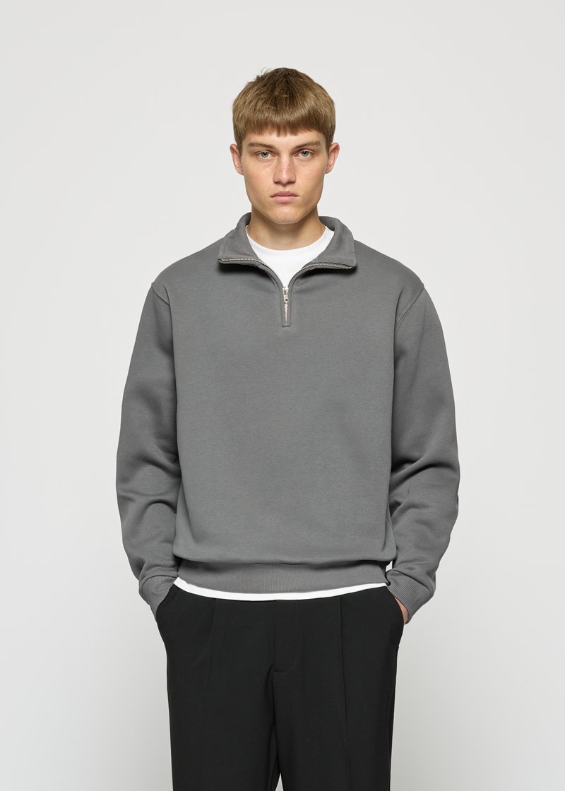 Jonge man met kort lichtbruin haar draagt een grijze sweatshirt met kwartrits, een wit shirt eronder en zwarte broek, staand met zijn handen in zijn zakken.