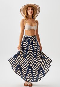 Falda larga con patrón de chevrones en azul marino y crema; tela ligera; cintura ajustada; combinada con un top de bikini neutro y un sombrero de paja de ala ancha.