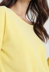 Gelbes, strukturiertes, locker sitzendes Oberteil mit weitem Ausschnitt und leicht herabgesetzten Schultern. Der Stoff wirkt atmungsaktiv und leicht.