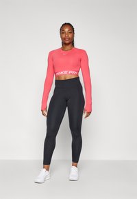 Nike Pro avkortet langermet topp i lys rosa med hvit logo, kombinert med høyt liv svarte leggings og hvite sportsko.