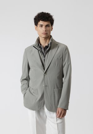 Hombre con cabello rizado que lleva una chaqueta y un blazer gris superpuestos sobre una camisa oscura, de pie contra un fondo claro y liso.