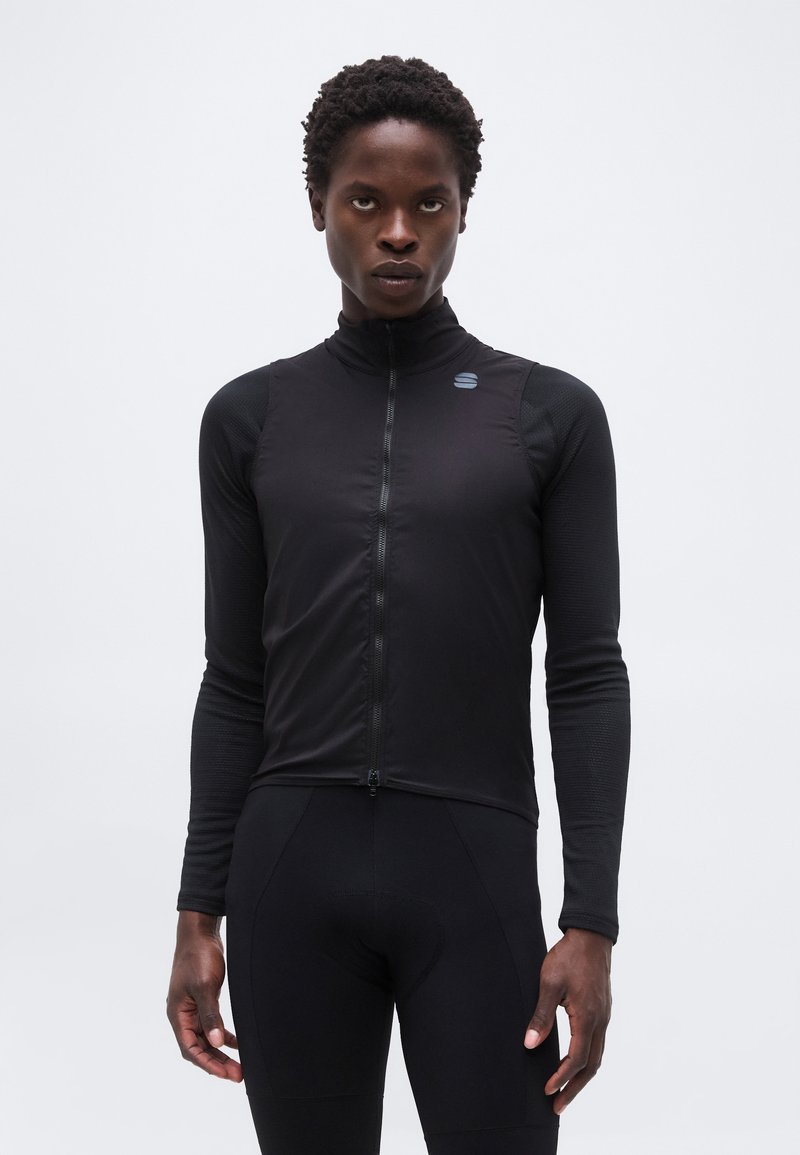 Λεπτός άνθρωπος που φοράει εφαρμοστό μαύρο ποδηλατικό μπλουζάκι με μακριά μανίκια, μπροστινό φερμουάρ και διακριτικό λογότυπο στο στήθος, στέκεται μπροστά σε απλό φόντο.