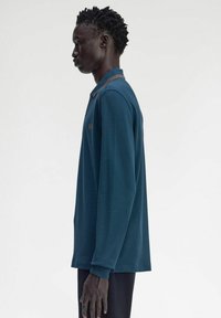 Polo shirt teal a maniche lunghe con colletto, caratterizzato da dettagli marroni e un logo ricamato. Il tessuto appare liscio e leggermente strutturato.