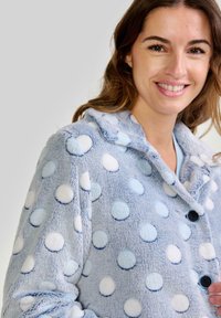 Chaqueta de forro polar azul claro con lunares blancos y azules claros, cierre de botón y diseño de cuello con reborde. Textura suave visible.