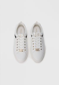 White sneakers with black side stripes, gold eyelets, and a decorative metal accent near laces. Smooth material and rounded toe design.