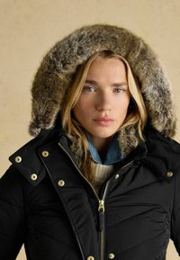 Melna izolēta jaka ar mākslīgā kažoka oderi kapucē. Iespējas: rāvējslēdzēja aizdare, zelta aksesuāri un izšūta raksts uz auduma.