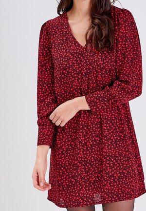 Femme portant une robe rouge et noire à manches longues avec un petit motif floral et un col en V, debout devant un fond clair uni.