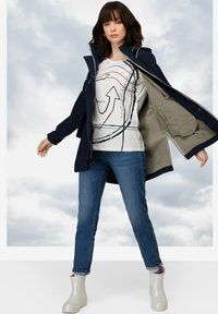 Chaqueta exterior impermeable de color azul marino con interior beige, combinada con una camiseta gráfica blanca, jeans azules y botas de tobillo blancas y brillantes.