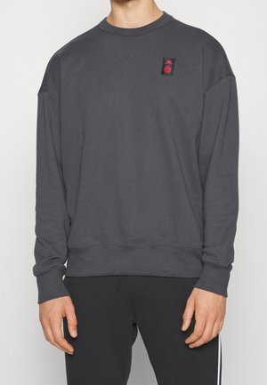 Personne portant un sweat-shirt à manches longues gris foncé avec un petit logo rouge Adidas sur la poitrine gauche et un pantalon noir avec une bande blanche.