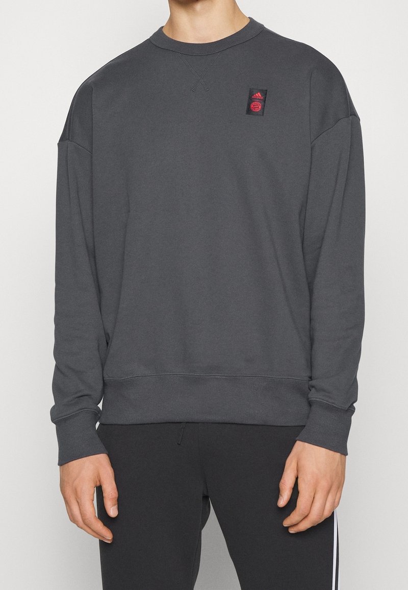 Personne portant un sweat-shirt à manches longues gris foncé avec un petit logo rouge Adidas sur la poitrine gauche et un pantalon noir avec une bande blanche.