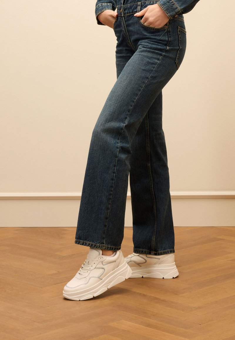 Jeans in denim a zampa d'elefante di colore blu scuro con orlo grezzo e cuciture laterali, abbinati a sneakers bianche con un design testurizzato e suola spessa.