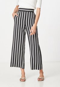 Žena nosící černobílé široké kalhoty s vertikálními pruhy, které mají zkrácenou délku, s rukama v kapsách a béžovými sandály s otevřenou špičkou na bílém pozadí.