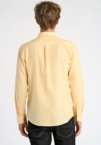 Heren langemouwen overhemd in lichtgeel, gemaakt van gestructureerde stof, met een achterste plooi en knoopmanchetten, gecombineerd met donkere denim jeans.