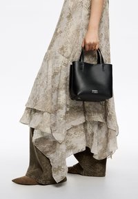 Zwart leren handtas met korte handvatten en een reliëflogo. Gecombineerd met een vloeiende, meerlagige, beige patroondress en olijfgroene broeken.