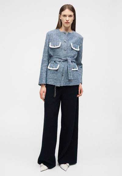 Γυναίκα που φοράει μπλε υφασμένο σακάκι με ζώνη και τσέπες με λευκή επένδυση, μαύρο παντελόνι με φαρδιά μπατζάκια και λευκά μυτερά παπούτσια, στεκόμενη μπροστά σε απλό φόντο.