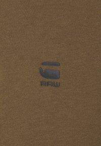 Tissu en coton marron avec une texture côtelée, présentant un petit logo bleu foncé de la marque "RAW" dans le coin inférieur droit.