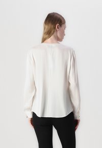 Blusa bianca a maniche lunghe con una texture liscia e setosa, scollo rotondo e una leggera arricciatura sulle spalle. Presenta una cucitura dritta sul retro.