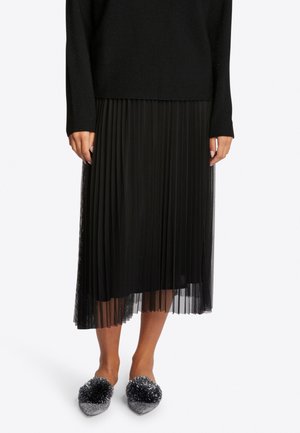 Modelo femenina con falda midi plisada negra, top de manga larga negro y zapatos planos plateados con purpurina y pompones negros, posando sobre fondo blanco.