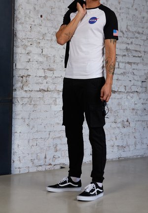 Mann trägt ein weißes und schwarzes NASA-T-Shirt, schwarze Cargohosen und schwarze Sneakers, steht vor einer weißen Ziegelwand und hält eine Sonnenbrille.