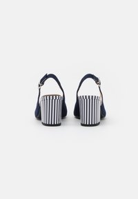 Sandali con tacco in suede blu navy con una fascia alla caviglia sul retro e un tacco a blocco a righe. Il tacco presenta righe verticali bianche e blu scuro alternate.