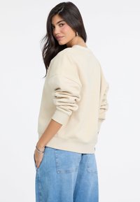 Beżowa bluza z opadającymi ramionami i ściągaczami, zestawiona z jasnoniebieskimi, szerokimi spodniami dżinsowymi. Miękka struktura, casualowy design, minimalne logo.