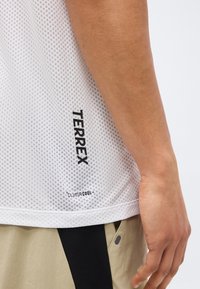 Κοντινό πλάνο του χεριού ενός ατόμου δίπλα σε λευκό αθλητικό πουκάμισο με δίχτυ με λογότυπα «TERREX» και «CLIMACOOL», συνδυασμένο με μπεζ και μαύρο παντελόνι.