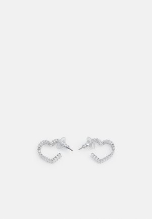 Boucles d'oreilles clous en forme de cœur ton argent avec cristaux clairs taillés en carré et attaches plastiques transparentes sur fond blanc.